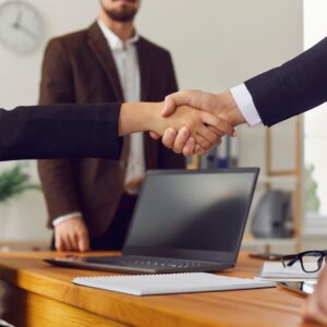Two business people shaking hands while a third looks on, all are wearing suits